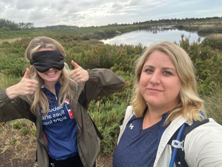 Rosie is wearing a blindfold and has two thumbs up and a huge smile. Kat is standing next to her and behind them there are green fields and a lake.