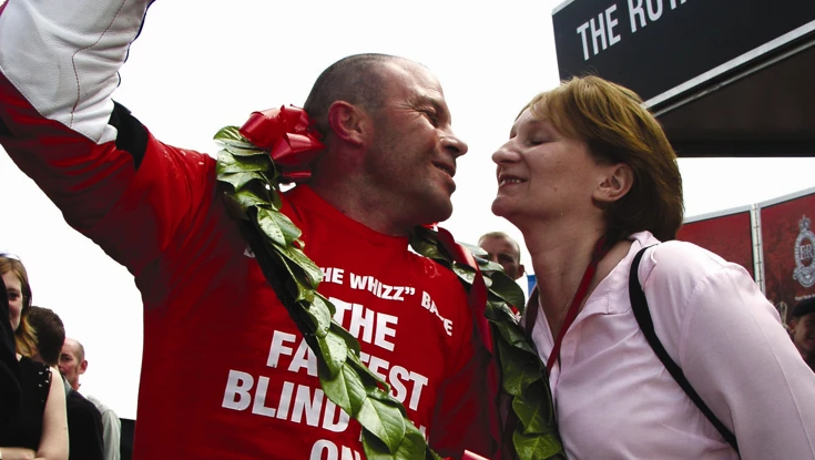 Blind veteran Billy, cheering in celebration while wearing motorcycle gear and holding a bottle of champagne as he leans in to kiss his wife