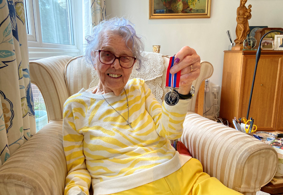 Blind veteran Gwynneth, sitting in her armchair at home, proudly holding up her WW2 medal