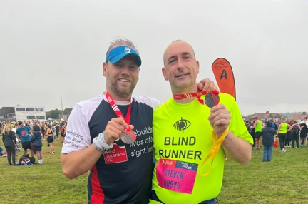 Blind veteran Steve and his guide runner Chris are holding up their medals to the camera. They are stood with their arms around one another's shoulders. Chris is wearing a Blind Veterans UK running top and Steve is wearing a bright yellow t-shirt with "blind runner" written on it.