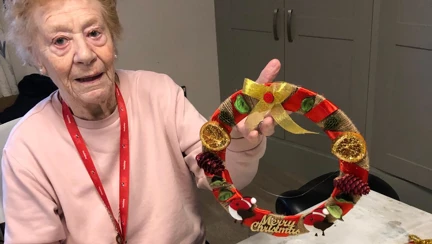 Blind veteran Daphne is holding up a wreath she decorated as she sits at a table covered in gold glitter