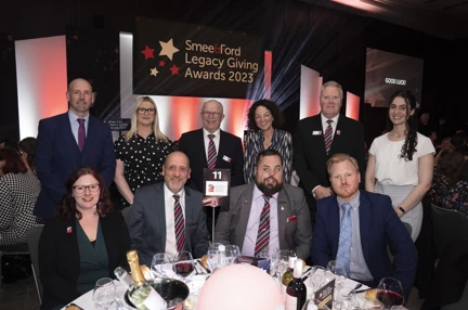 A group of Blind Veterans UK staff, sitting and standing at a round table at an awards ceremony, with a banner behind them that reads: Smee & Ford Legacy Giving Awards 2023