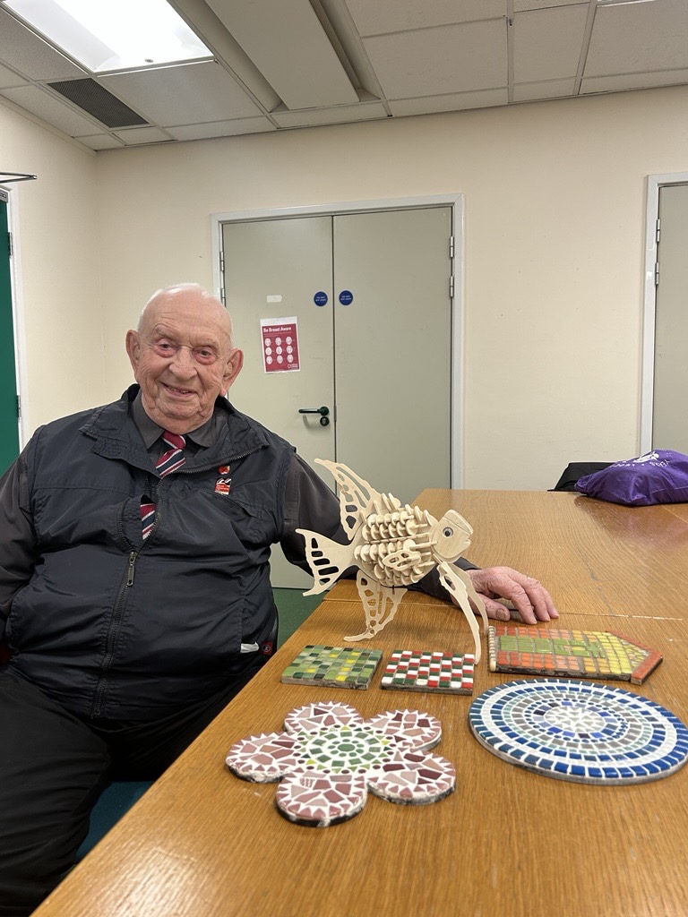 Glynne is seated at a table smiling, on the table next to him are a selection of wooden models and mosaics he's worked on.