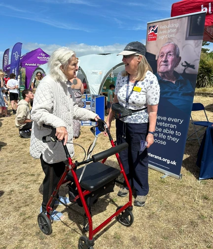 Blind veteran Win is leaning on a walker as she talks to a volunteer standing next to a Blind Veterans UK banner