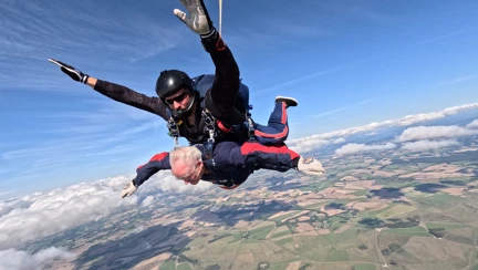 Danny during the parachute jump. The parachute is open and Danny is gliding towards the ground. The sky is blue and below are lots of green fields.