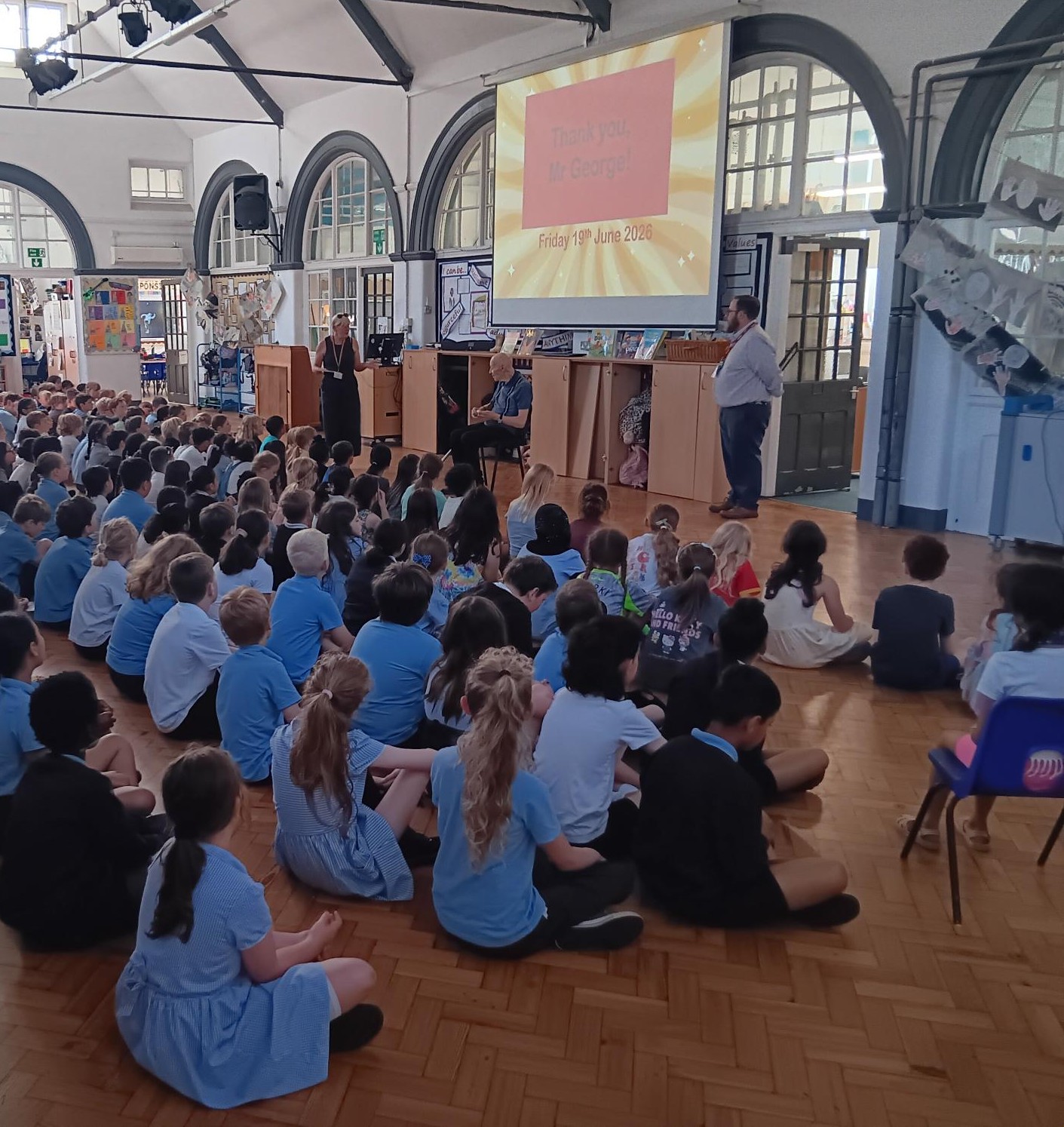 Dennis sits on a chair in assembly in front of a large group of children