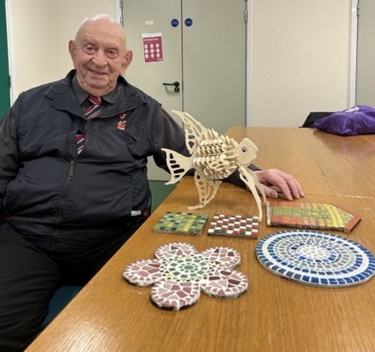 Glynne sat next to his creative projects – A wooden model of an angelfish, surrounded by colourful mosaics
