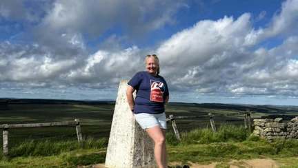 Janine is leaning against a marker point on the trail. She is smiling and wearing her Blind Veterans UK t-shirt