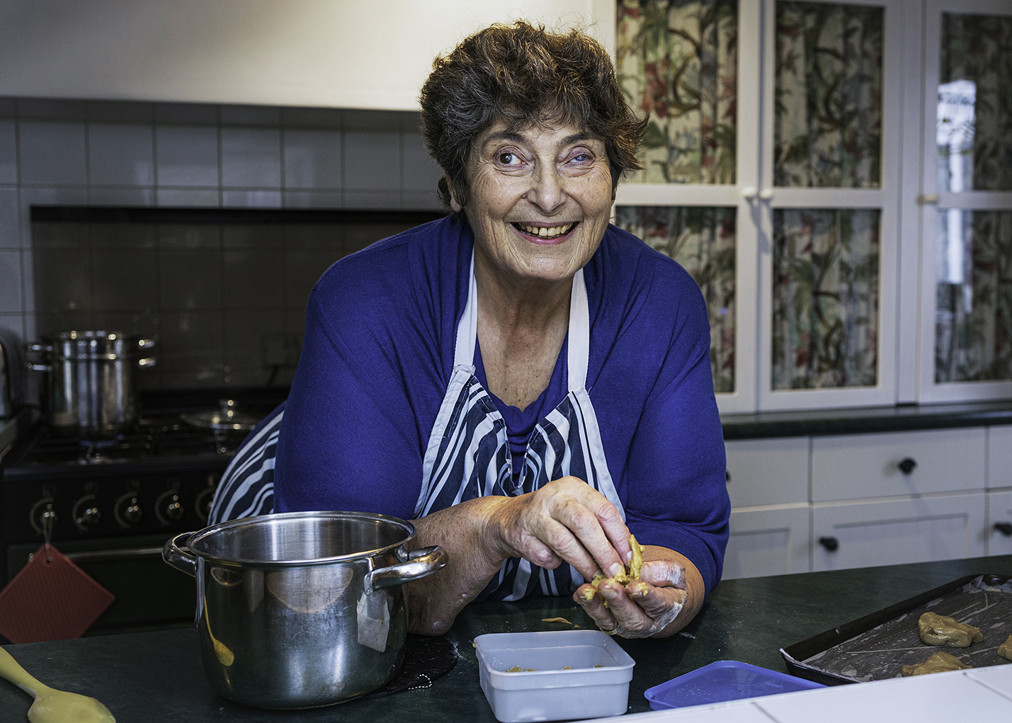 Penny leaning on her kitchen sideboard smiling and looking at the camera. Her oven behind her and a bowl beside her on the left and a baking tray on the right. 