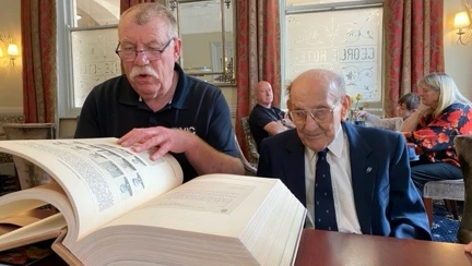Matt is sitting at a table as a man next to him turns the page on a giant book