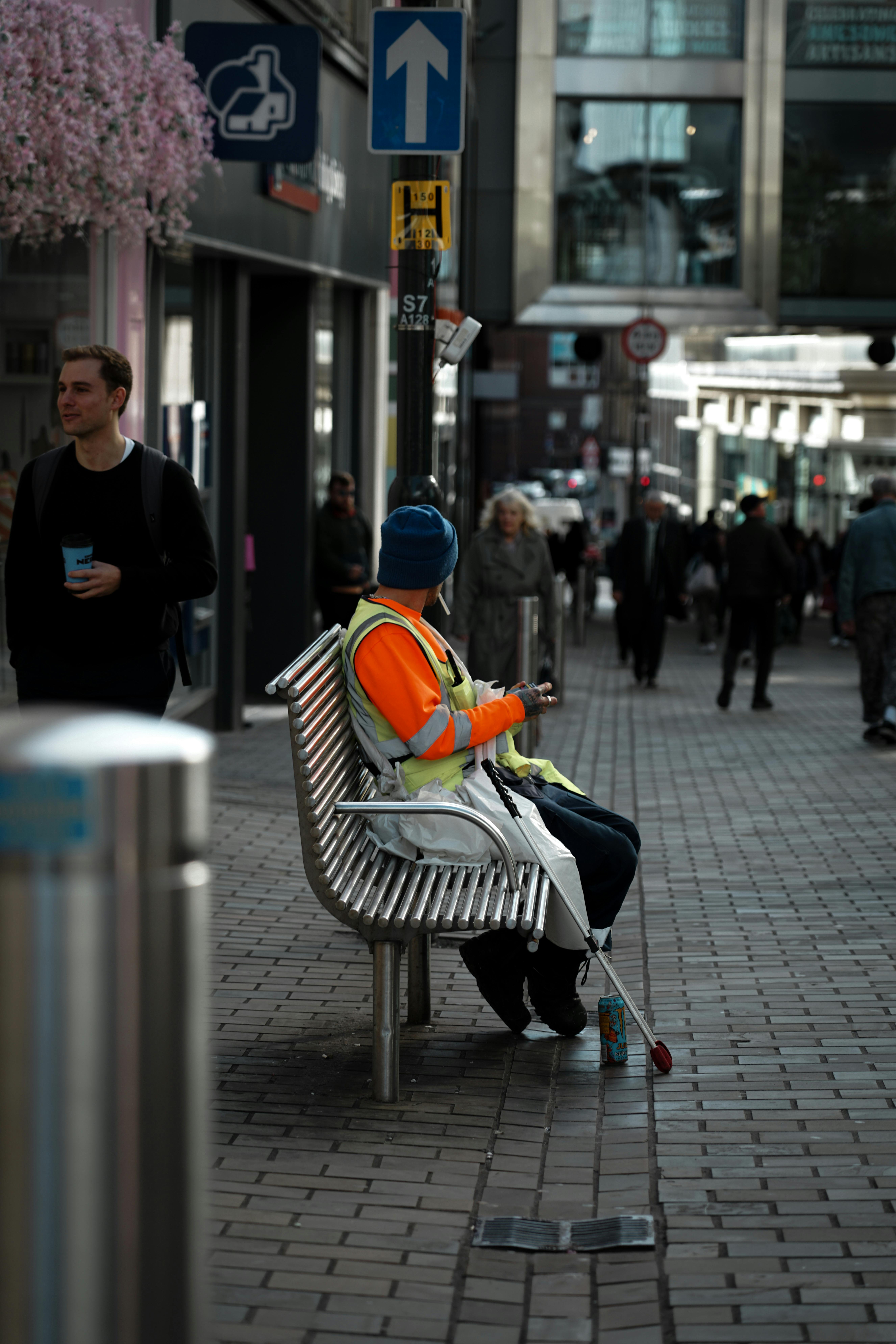 A person wearing a high-visibility vest and hat sits on a metal bench on a city street, holding a white cane, with people walking along the pavement in the background.