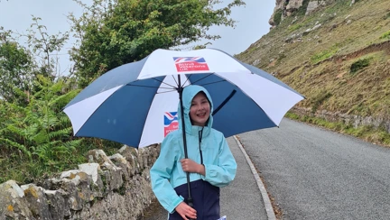 A photo of Emmie, wearing a a hooded rain coat and holding a Blind Veterans UK umbrella, walking along a curved path