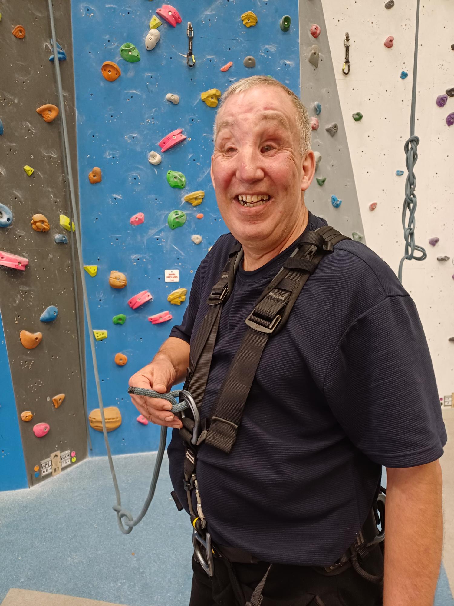 Carl is roped up and smiles while standing in front of a large climbing wall