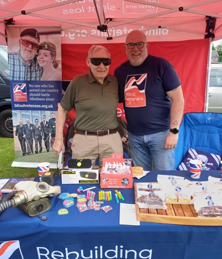 A blind veteran wearing dark glasses with a Blind Veterans UK member of staff, who is wearing a branded t-shirt. They stand, both smiling, at a decorated Armed Forces Day stall.