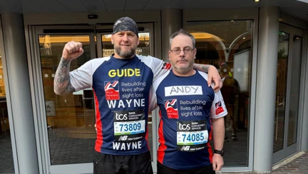 Andy and Wayne in their Blind Veterans UK T-shirts standing with their arms around each other. Wayne has 'Guide' written on his T-shirt and Andy is wearing Union Jack socks