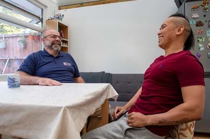 Blind veteran Ken and his support worker sitting at a table laughing together in Ken's home