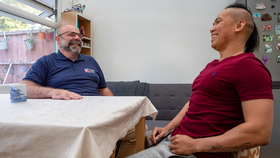 Blind veteran Ken and his support worker sitting at a table laughing together in Ken's home