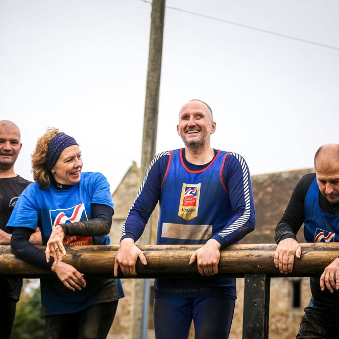 A group of four runners in Blind Veterans UK vests, muddy and smiling as they rest after finishing their race