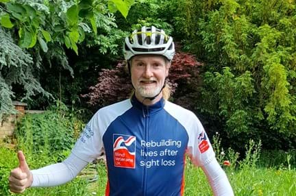 Keith stood with his bike wearing his helmet and a Blind Veterans UK cycling top. One hand is holding the bike up and the other is doing a thumbs up