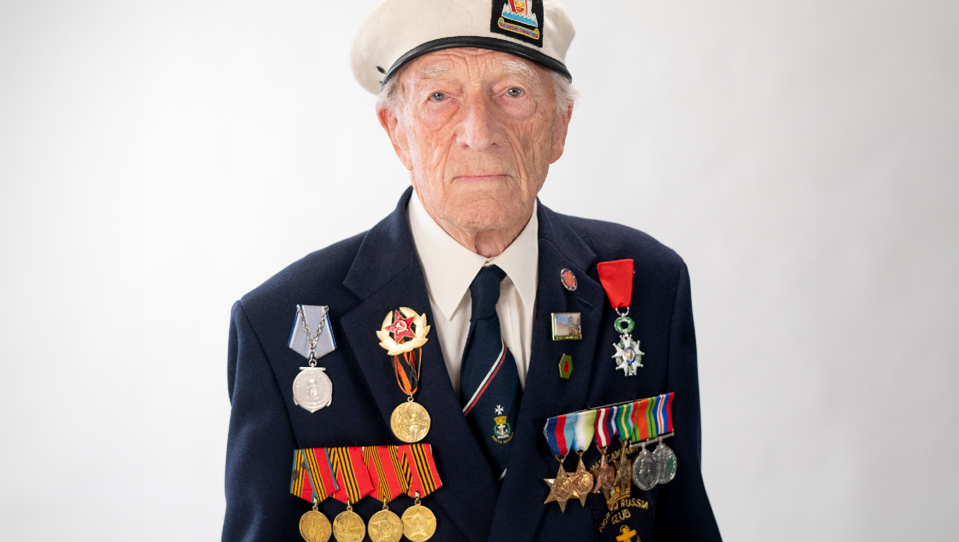 Alec is wearing a navy blazer, cream beret, medals, and looks proudly into the camera