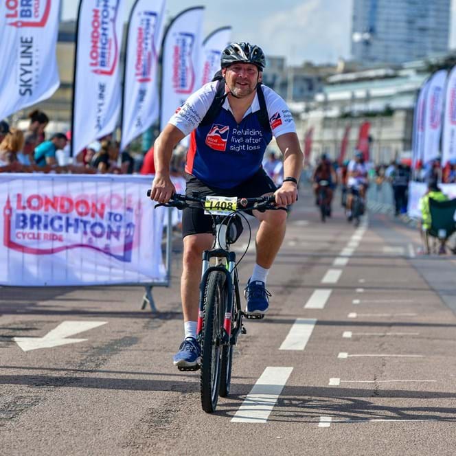 Gary crossing the finish line of the race on his bike wearing a Blind Veterans UK t shirt.