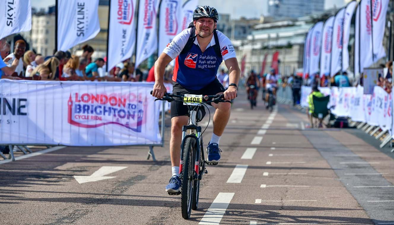Gary crossing the finish line of the race on his bike wearing a Blind Veterans UK t shirt.