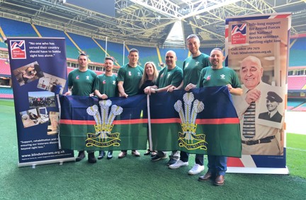 A group of supporters in green t-shirts, standing in a rugby stadium, between two Blind Veterans UK pull up banners, holding up a Royal Welsh banner,