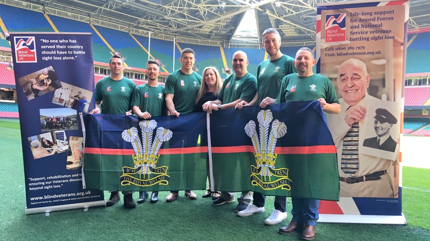 A group of supporters in green t-shirts, standing in a rugby stadium, between two Blind Veterans UK pull up banners, holding up a Royal Welsh banner,