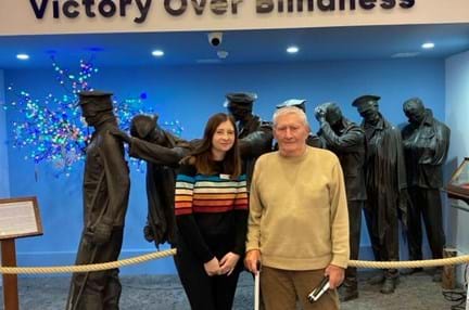 Stephanie and Mike are stood together in front of the 'Victory over Blindness' sculpture depicting a group of soldiers from the First World War leading one another away from the battlefield. Mike is holding hi white cane.