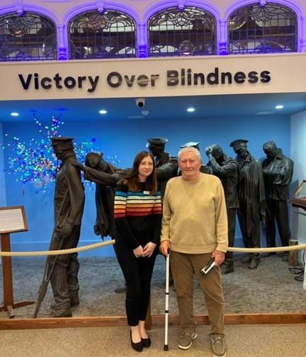 Stephanie and Mike are stood together in front of the 'Victory over Blindness' sculpture depicting a group of soldiers from the First World War leading one another away from the battlefield. Mike is holding hi white cane.