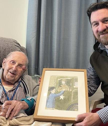 Ken and Tim are sat next to each other smiling and Tim is holding a framed illustration that he's created of Ken from his D-Day memories. In the picture a young Ken is sat at the steering wheel of his lorry leaning out the window as he talks to a Frenchman.
