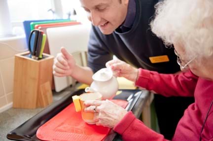 Photo of blind veteran pouring a cup of tea with her liquid level indicator and staff member leaning over her shoulder to help