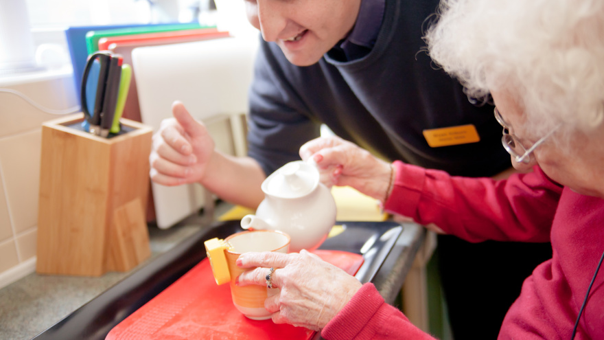 A blind veteran pouring a cup of tea, using a liquid level indicator, and a support worker leaning over her shoulder to help.