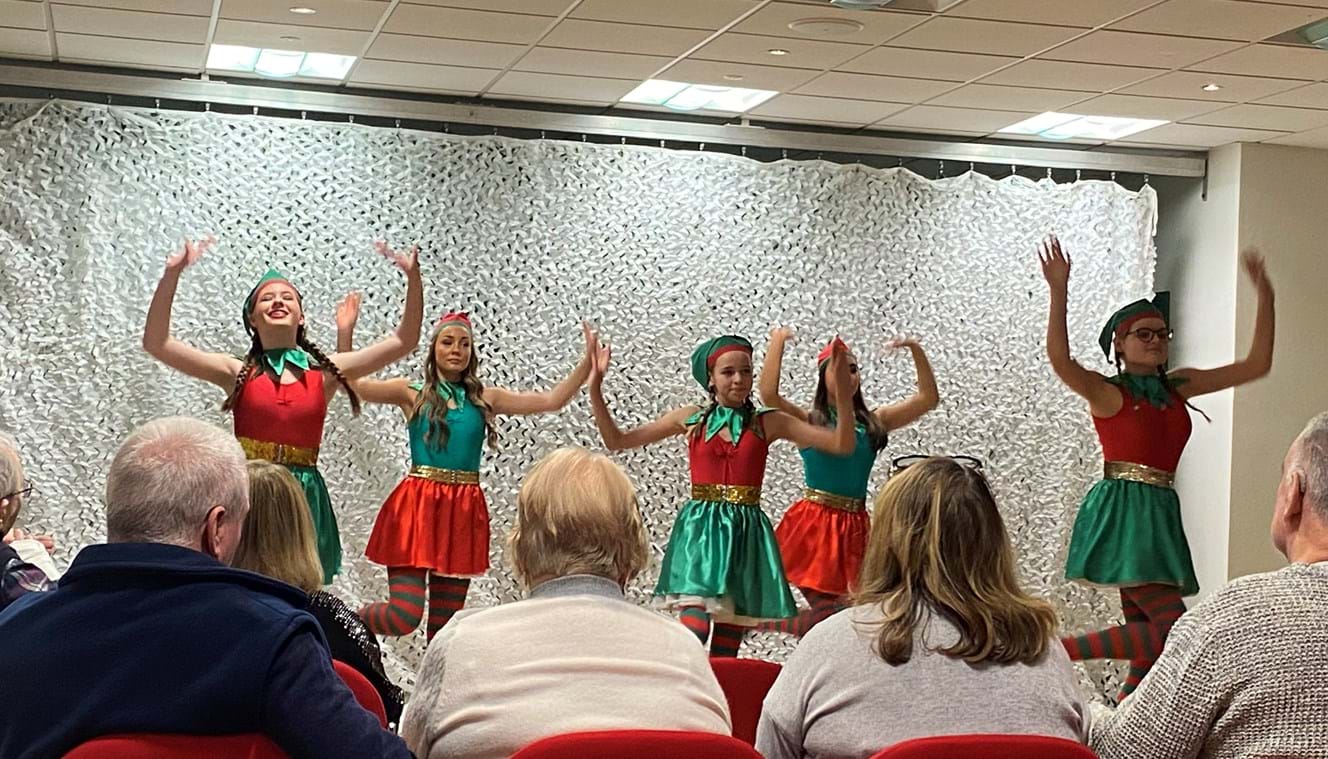 Five girls on stage dressed as elves tap dancing for an audience of blind veterans