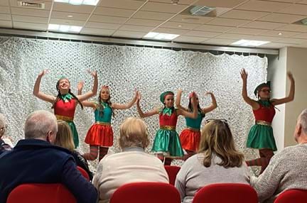 Five girls on stage dressed as elves tap dancing for an audience of blind veterans
