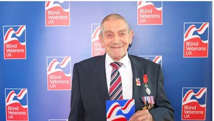 Blind veteran Eddie is wearing a suit and medals. He holds his Founder's Day award and looks into the camera. There is a Blind Veterans UK branded backdrop.