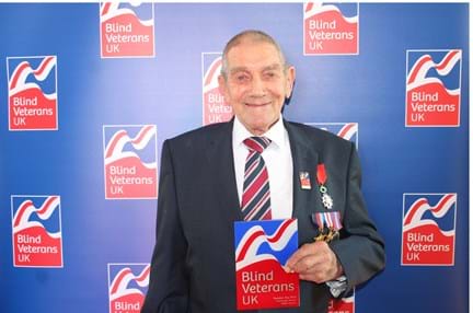Blind veteran Eddie is wearing a suit and medals. He holds his Founder's Day award and looks into the camera. There is a Blind Veterans UK branded backdrop.