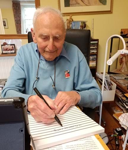 Blind veteran John, sitting at a desk with a large notepad, pen in hand, writing his memoirs