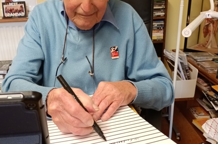 Blind veteran John, sitting at a desk with a large notepad, pen in hand, writing his memoirs