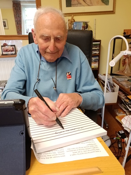 Blind veteran John, sitting at a desk with a large notepad, pen in hand, writing his memoirs
