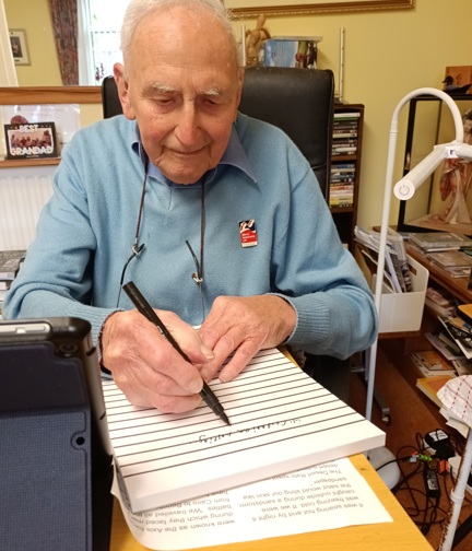 Blind veteran John, sitting at a desk with a large notepad, pen in hand, writing his memoirs