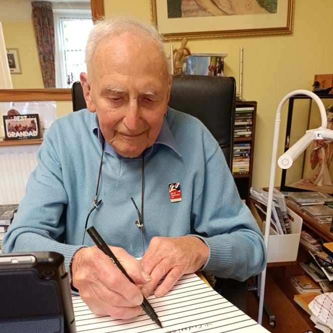 Blind veteran John, sitting at a desk with a large notepad, pen in hand, writing his memoirs
