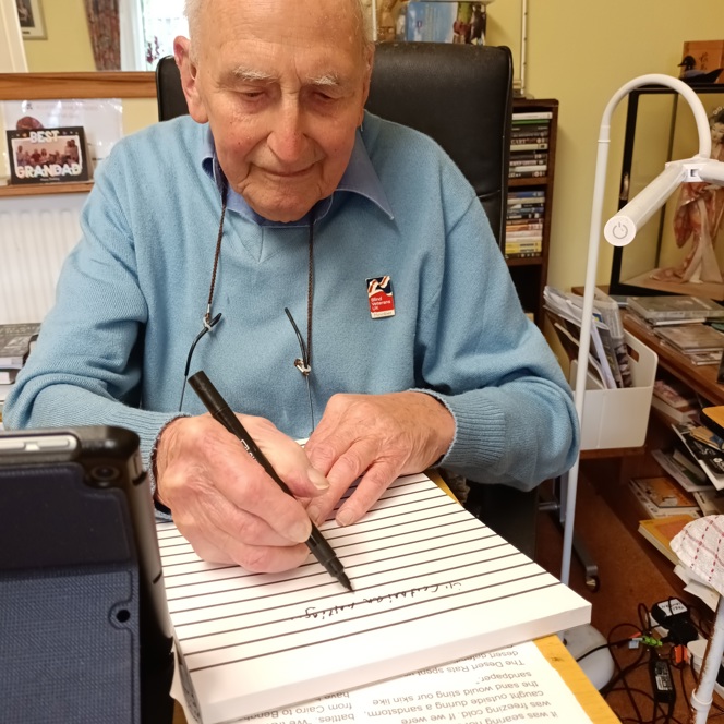 Blind veteran John, sitting at a desk with a large notepad, pen in hand, writing his memoirs