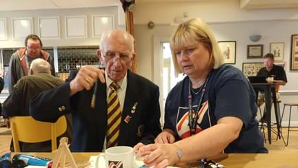 Bob is holding a paint brush sat at a table next to his Community Support Worker