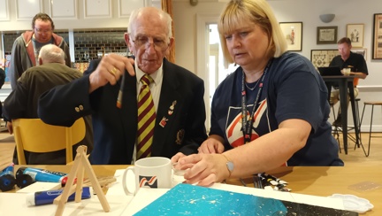 Bob is holding a paint brush sat at a table next to his Community Support Worker