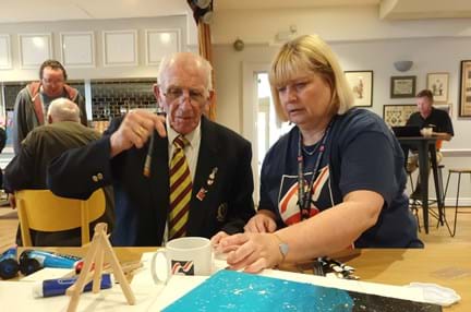 Bob is holding a paint brush sat at a table next to his Community Support Worker