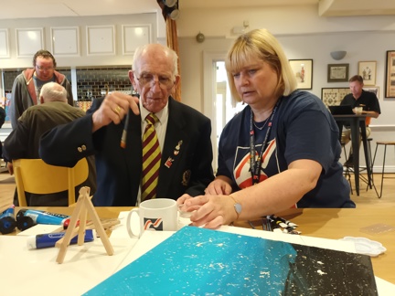Bob is holding a paint brush sat at a table next to his Community Support Worker