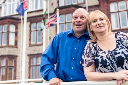 Blind veteran Billy with his arm around his wife, smiling as they stand outside our Centre of wellbeing in Llandudno