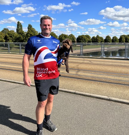 Simon stood in front of a canal in the sunshine wearing his Blind Veterans UK top and holding his dog
