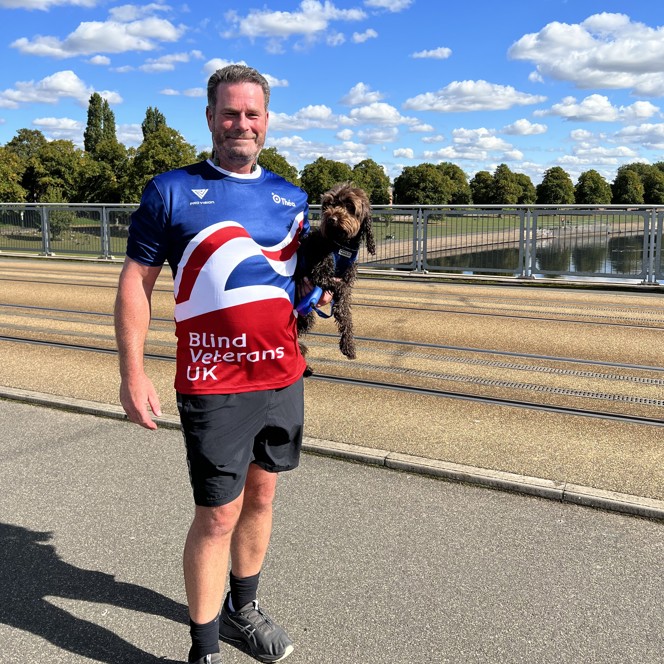 Simon stood in front of a canal in the sunshine wearing his Blind Veterans UK top and holding his dog